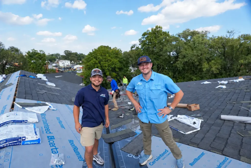 Two Jade Roofing & Construction team members in company polos smiling on a roof mid-replacement, with new architectural shingles and synthetic underlayment behind them